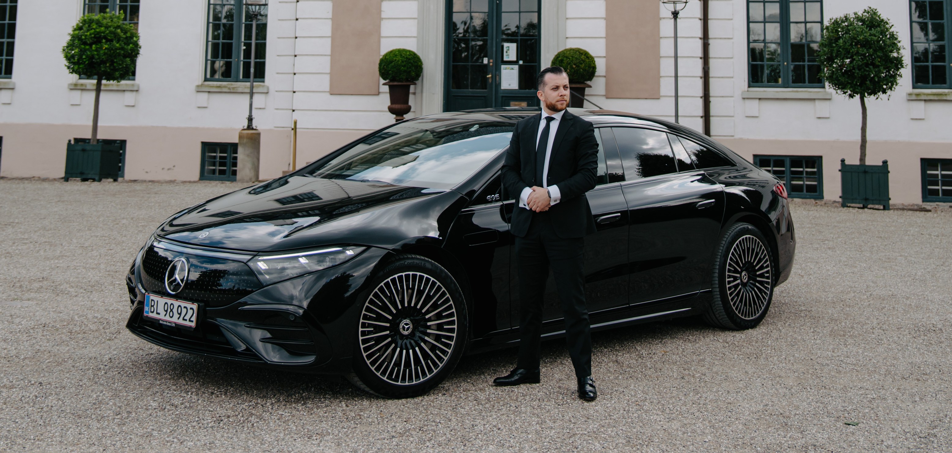 LuxLimo chauffeur in a black suit standing beside a Mercedes-Benz EQS outside a Danish manor house