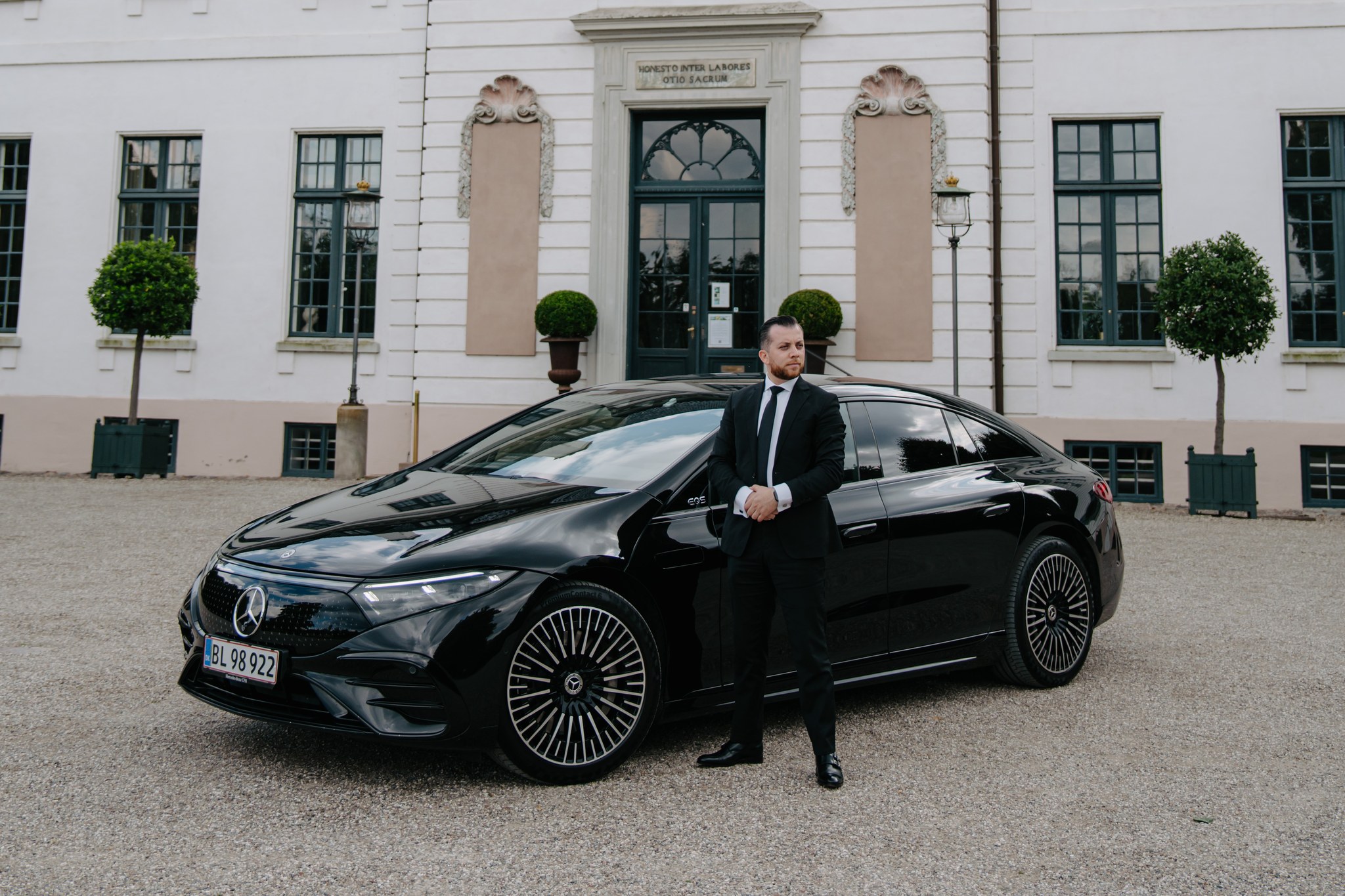 LuxLimo chauffeur in a black suit standing beside a Mercedes-Benz EQS outside a Danish manor house