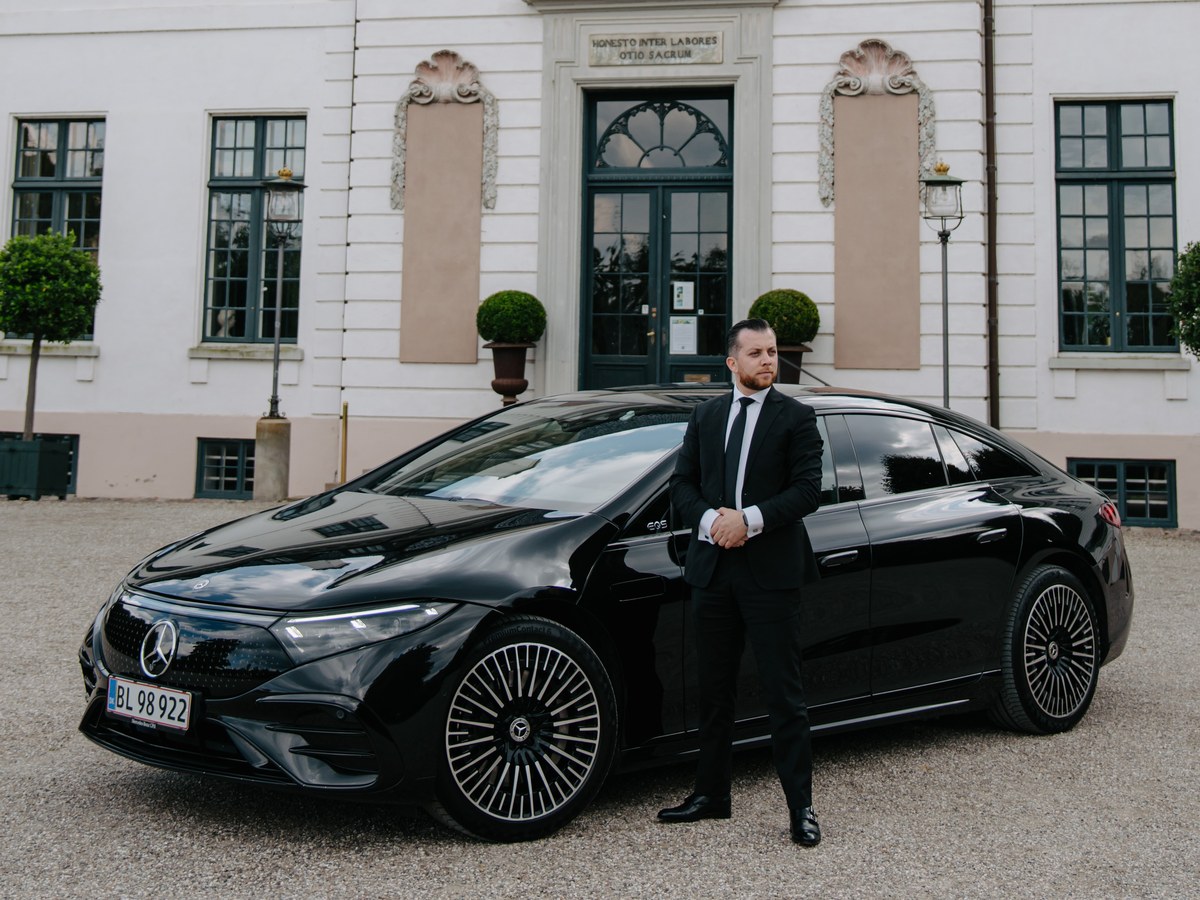 LuxLimo chauffeur in a black suit standing beside a Mercedes-Benz EQS outside a Danish manor house