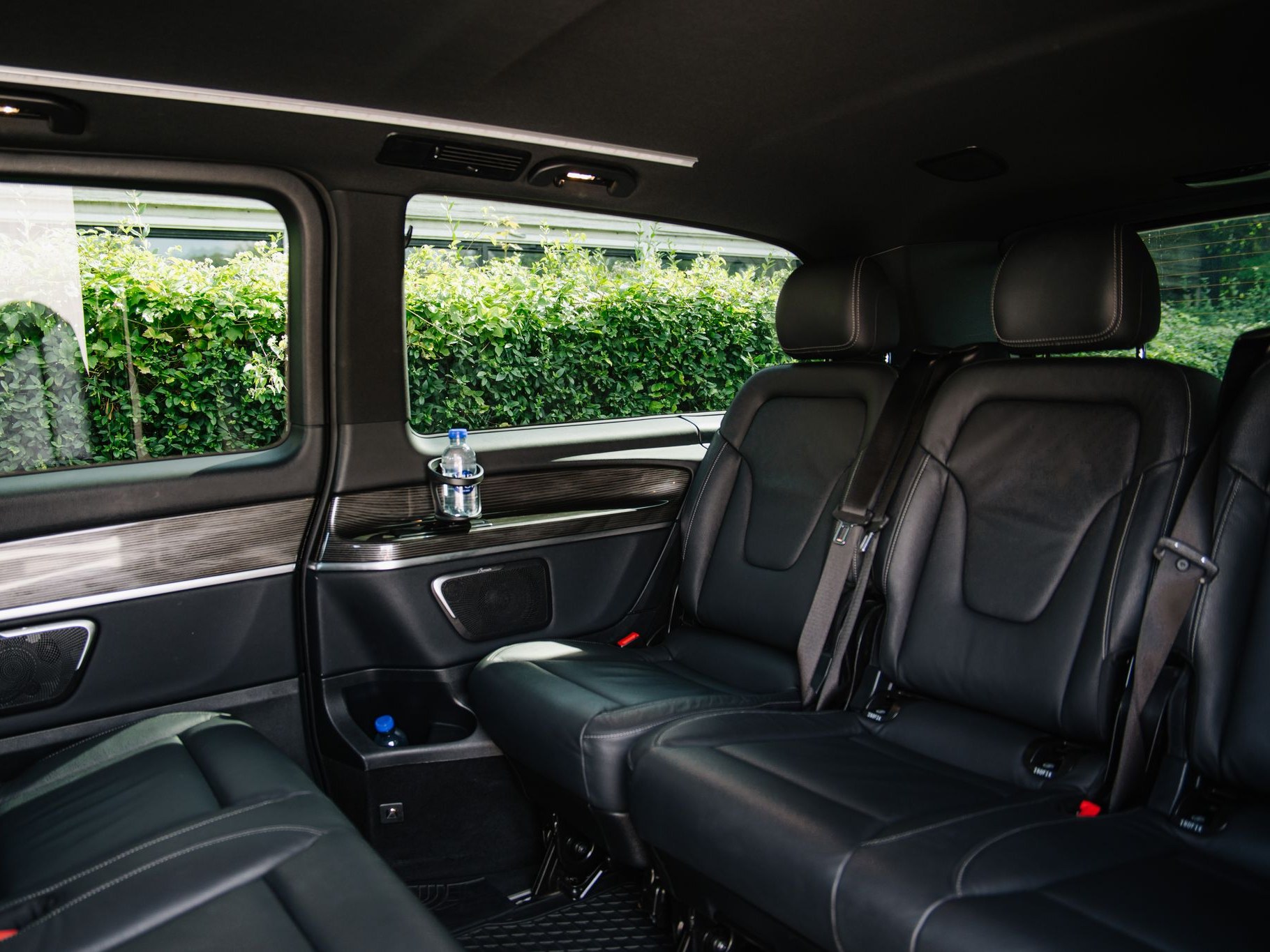 The rear cabin of the LuxLimo Mercedes-Benz EQV: black leather seats with headrests, a bottle of water on the door ledge and the hedge outside the window