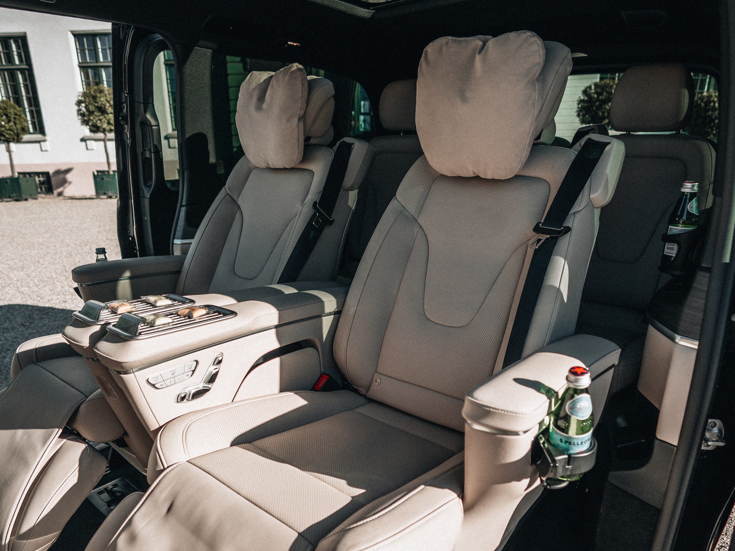 Inside the LuxLimo Mercedes-Benz EQV: two light leather lounge seats with headrests, a table between them and a bottle of water, seen through the open sliding door