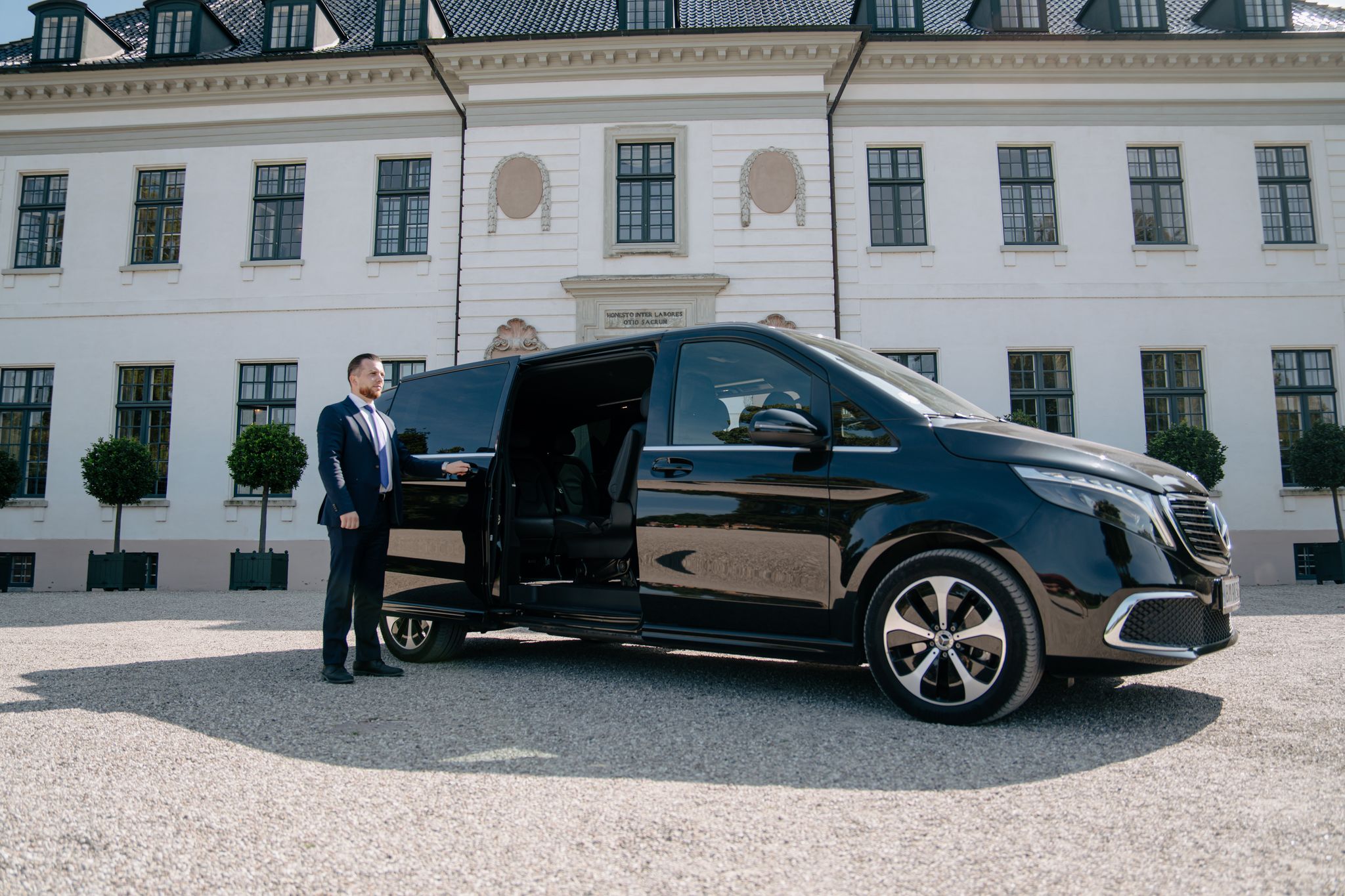 LuxLimo chauffeur at the open sliding door of the Mercedes-Benz EQV, XL, at a Danish manor house